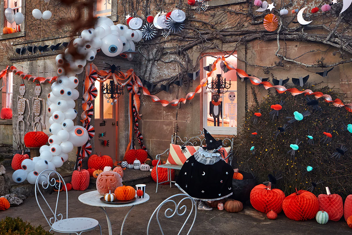 Meri Meri Akcesoria na imprezę o tematyce Halloween wystawione przed rustykalnym domem, w tym pastelowe papierowe girlandy, balonowy łuk w kształcie gałki ocznej i dynie o strukturze plastra miodu